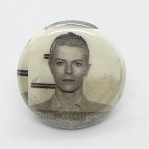 Puede incluir: Un botón en blanco y negro con una foto de carnet de David Bowie. El botón tiene el texto "Police Department Rochester, NY" impreso en la parte inferior.