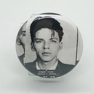 Puede incluir: Botón en blanco y negro estilo foto policial con un joven de pelo oscuro. El texto "VERNON COUNTY SHERIFF'S OFFICE" y "67 30" son visibles en la parte inferior del botón.