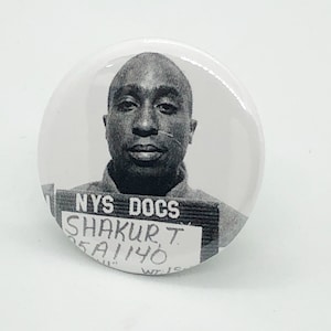 Puede incluir: Foto en blanco y negro estilo ficha policial de un hombre con el texto "NYS DOCS" y "SHAKUR T. 05A1140" sobre un fondo blanco.