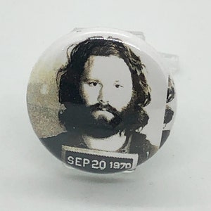 Puede incluir: Botón de foto en blanco y negro con un hombre de pelo largo y barba. La foto es un retrato policial con la fecha "SEP 20 1970" impresa debajo.