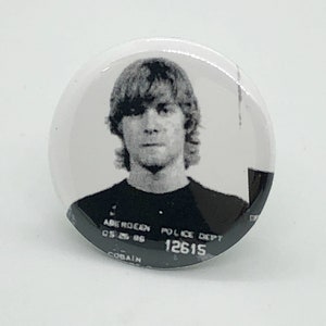 Puede incluir: Botón de foto en blanco y negro con un joven con el pelo corto y ondulado. La foto es un retrato policial con el texto "Police Dept. 12615" y "Kurt Cobain" visible.
