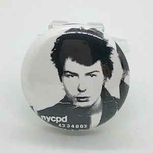 Puede incluir: Botón blanco y negro con una foto de una persona con el pelo corto y oscuro. El botón tiene el texto "nycpd 4334003" impreso en él.