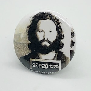 Puede incluir: Botón de foto en blanco y negro con una fotografía de ficha policial de Jim Morrison con el texto "SEP 20 1970" y "CITY DEPT. - DADE" debajo.