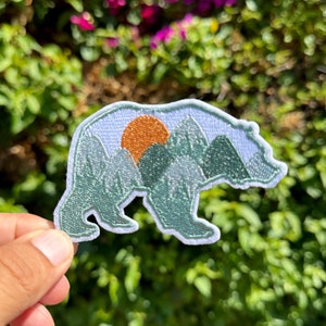 May include: A white embroidered patch of a bear silhouette with a mountain range and a setting sun inside the bear's body. The mountains are green and the sun is orange.