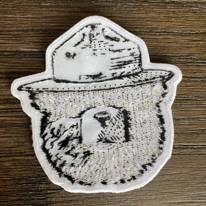May include: White embroidered patch with a black and white image of a bear wearing a ranger hat. The bear's face is visible and the hat has a brim.