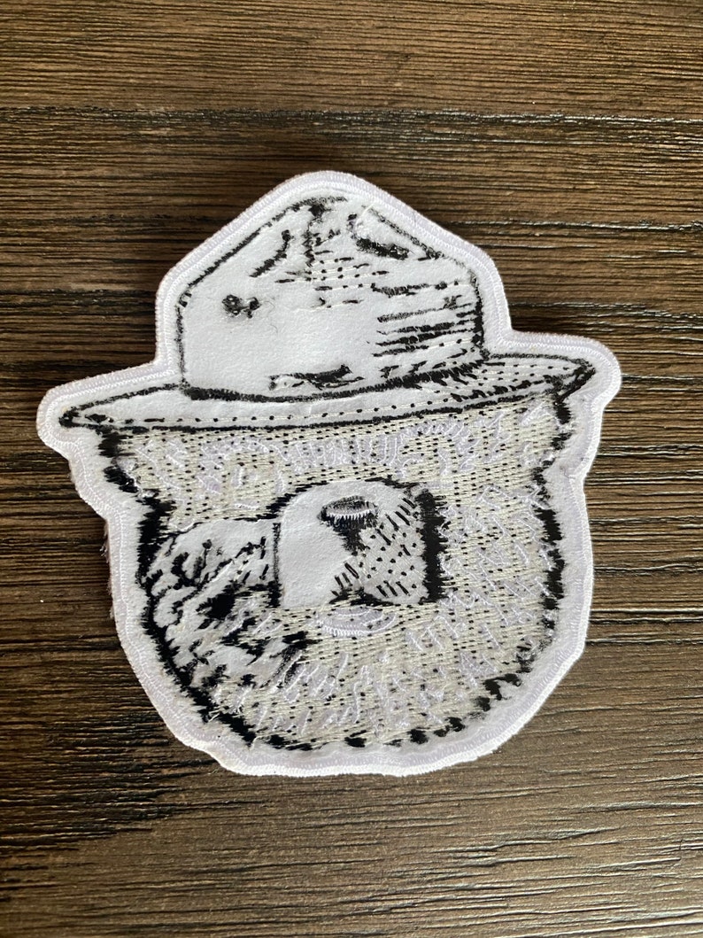 May include: White embroidered patch with a black and white image of a bear wearing a ranger hat. The bear's face is visible and the hat has a brim.