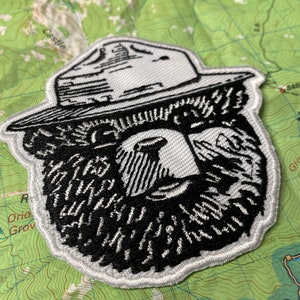 May include: A white embroidered patch with a black and white illustration of a bear wearing a ranger hat. The bear has a thick beard and a serious expression.