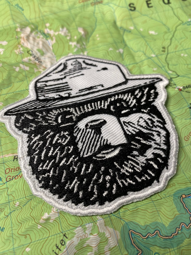 May include: A white embroidered patch with a black and white illustration of a bear wearing a ranger hat. The bear has a thick beard and a serious expression.