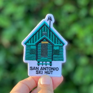 Puede incluir: Un parche bordado blanco con un diseño de cabaña verde y el texto "San Antonio Ski Hut".