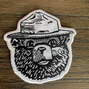 May include: White embroidered patch of a bear wearing a ranger hat. The bear has a black beard and eyes. The hat is black with white stitching.