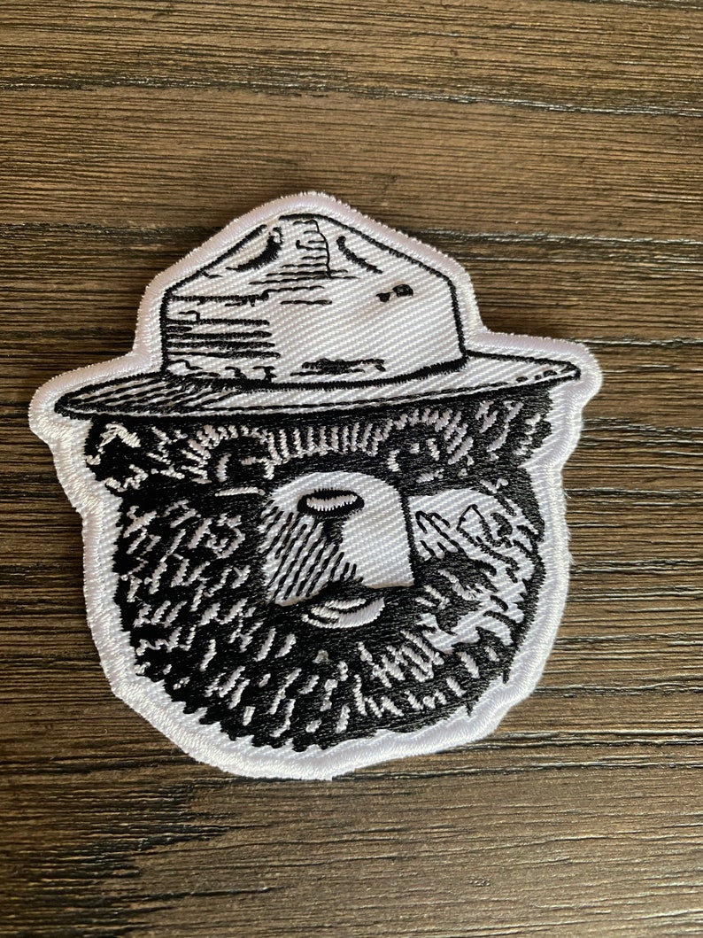 May include: White embroidered patch of a bear wearing a ranger hat. The bear has a black beard and eyes. The hat is black with white stitching.