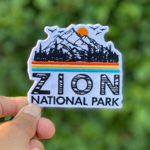 Puede incluir: Parche blanco bordado con una escena de montaña en blanco y negro, un sol poniente y el texto "ZION NATIONAL PARK".