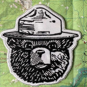 May include: Black and white embroidered patch of a bear wearing a ranger hat. The bear has a thick beard and a serious expression.