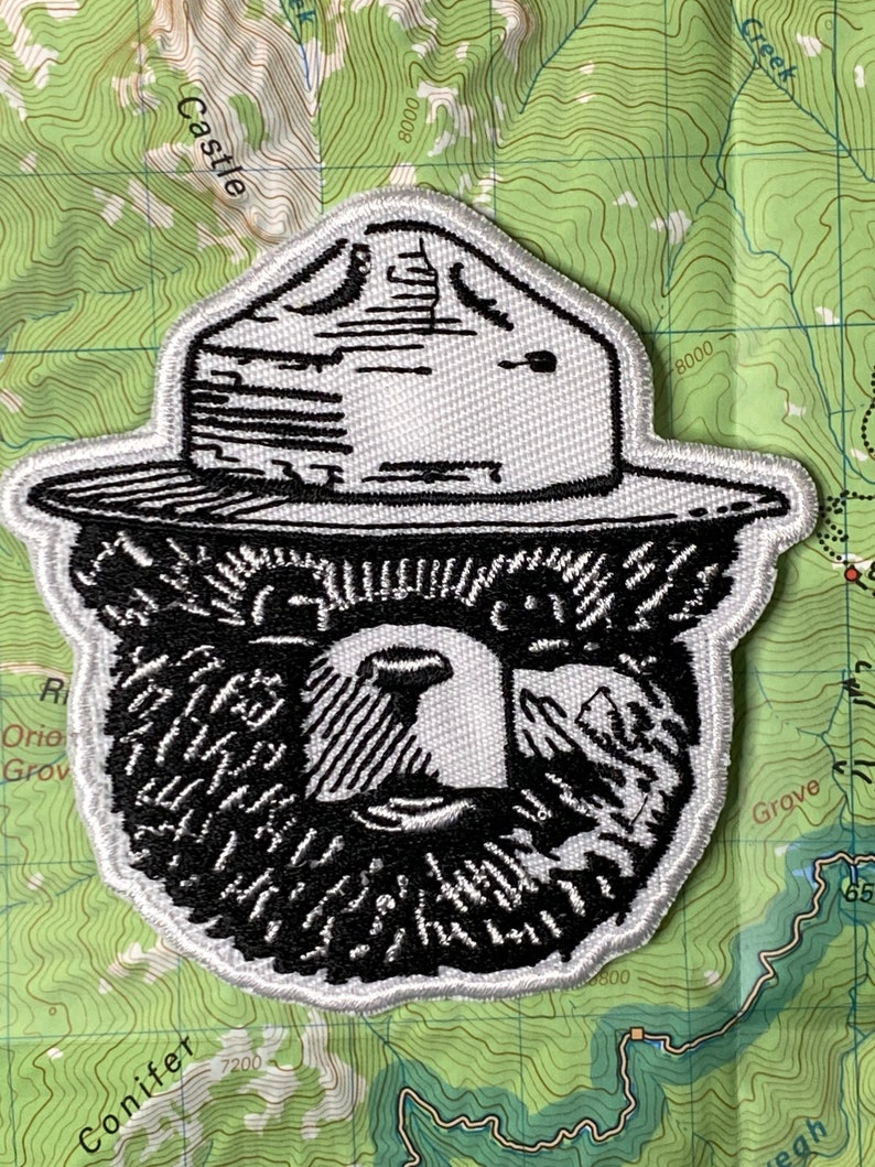 May include: Black and white embroidered patch of a bear wearing a ranger hat. The bear has a thick beard and a serious expression.