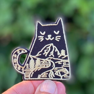 May include: Black embroidered iron-on patch of a cat with a mountain landscape inside its silhouette. The cat has a white outline and white eyes. The mountain landscape is also white.