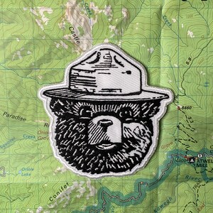 May include: Black and white embroidered patch of a bear wearing a ranger hat. The patch is on a green background with topographic lines.