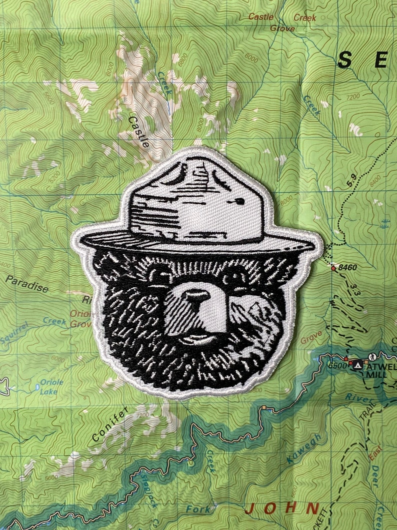 May include: Black and white embroidered patch of a bear wearing a ranger hat. The patch is on a green background with topographic lines.
