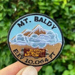 May include: A round, brown embroidered patch with a blue sky background. The patch features a mountain range with a white cloud above it. The text "MT. BALDY" is embroidered in black around the top of the patch. The text "ELEV. 10,064 FT" is embroidered in black around the bottom of the patch. A brown rectangular sign with the text "SAN ANTONIO MTN. VALLEY ELEV. 10,064" is embroidered in yellow on the patch.