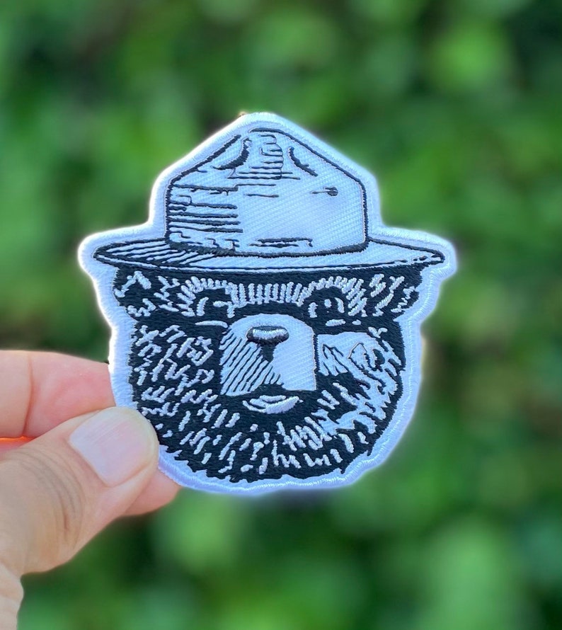 May include: White embroidered patch of a bear wearing a ranger hat. The bear's face is detailed with a black outline and black fur.