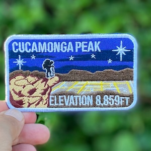 May include: A blue and white embroidered patch with the text "CUCA MONGA PEAK ELEVATION 8,859FT" and a depiction of a mountain peak with a star in the sky.