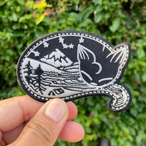 Peut inclure: Un patch brodé noir et blanc représentant un chat dormant sur une montagne avec des étoiles dans le ciel. Le chat est délimité en blanc et la montagne est ombragée en blanc.