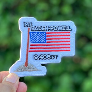 May include: Embroidered patch featuring the American flag with red and white stripes and a blue field of stars. The patch reads "MT. BADEN-POWELL" and "9,406 FT" in black text. The patch has a white border and a brown flagpole.