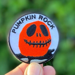 May include: A white and orange embroidered patch with the text "Pumpkin Rock" in black lettering. The patch features a cartoon pumpkin with a black face and a stitched smile.