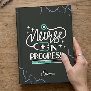 May include: A black notebook with white text that reads "Nurse In Progress" with a stethoscope graphic. The notebook has a marble-like design and the name "Sienna" at the bottom. The notebook is being held by a person.