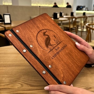 May include: A wooden menu with a dark stain, featuring a heron illustration and the text "BAR L'EFFERVESCENCE". The menu is held open, revealing a black spine and silver rivets. The background shows a restaurant setting.