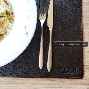 May include: A brown leather placemat with a stitched border and the text "SAFYANU LEATHER GOODS" embossed in the lower right corner. The text "Your logo can be placed here" is in a white box in the upper right corner.