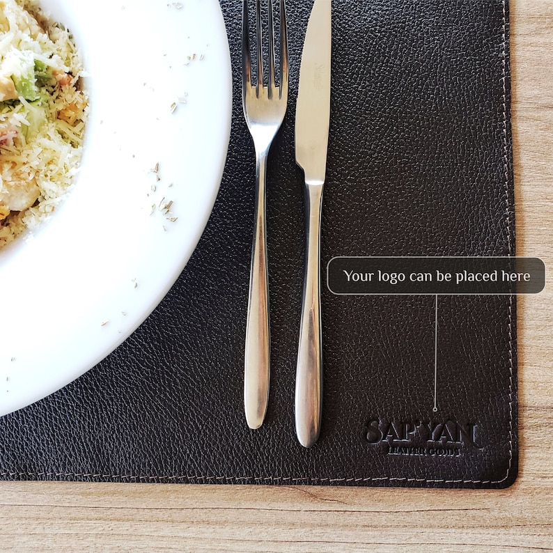 May include: A brown leather placemat with a stitched border and the text "SAFYANU LEATHER GOODS" embossed in the lower right corner. The text "Your logo can be placed here" is in a white box in the upper right corner.