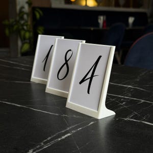 May include: Three white table number holders with the numbers 1, 8, and 4 in black script. The holders are on a black marble table. The background is blurred, showing a restaurant setting.