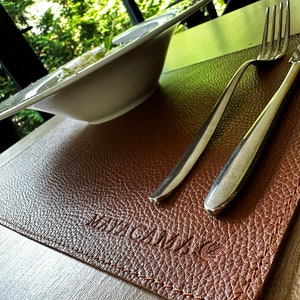 May include: Brown leather placemat with a stitched edge and the word "MAGNUM" embossed in the center. A silver fork and knife are placed on the mat.