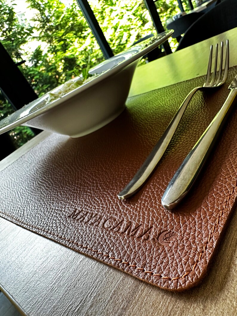 May include: Brown leather placemat with a stitched edge and the word "MAGNUM" embossed in the center. A silver fork and knife are placed on the mat.