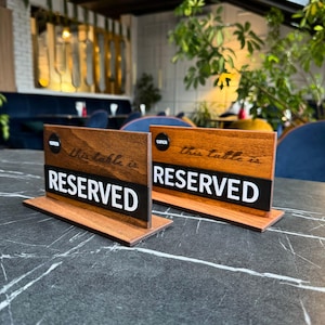 May include: Two wooden table signs with the word "RESERVED" in large white letters on a black stripe. The signs have a brown wooden base and top, with the text "this table is" in cursive. The word "LUNCH" is in a circle on the top left.