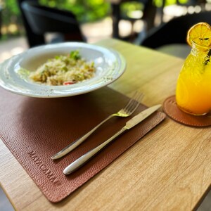 May include: A brown leather placemat with the word "MACA" printed on it. A white plate with a portion of pasta sits on the placemat. A fork and knife are on the placemat. A glass of orange juice with a slice of orange sits on a brown leather coaster.