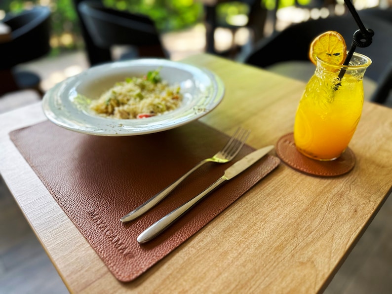 May include: A brown leather placemat with the word "MACA" printed on it. A white plate with a portion of pasta sits on the placemat. A fork and knife are on the placemat. A glass of orange juice with a slice of orange sits on a brown leather coaster.