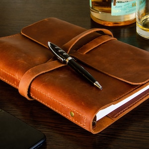 May include: A brown leather-bound journal with a pen resting on top. The journal is closed and has a strap that wraps around it. The journal is on a wooden surface.