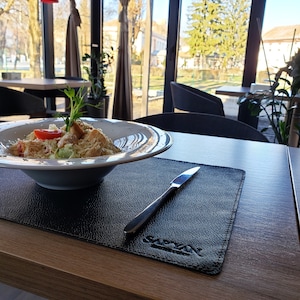 May include: A white bowl filled with a Caesar salad with croutons, lettuce, parmesan cheese, and a tomato. The bowl is on a black leather placemat with a silver knife and the word "SARAY" printed on the mat. The table is wooden.