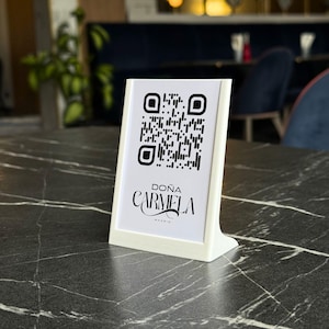 May include: A white table tent with a QR code and the text "DOÑA CARMELA MADRID" on a black marble table. The tent is angled towards the viewer, showcasing the restaurant's branding and a digital access point.