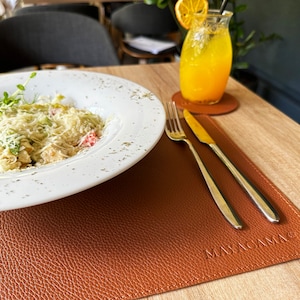 May include: A white plate with a salad topped with grated cheese, tomatoes, and a sprig of greenery. The plate is on a brown leather placemat with the text "MAYACAMA" embossed on it. A fork and knife are on the placemat. A glass of orange juice with a slice of orange is on a coaster to the right of the plate.