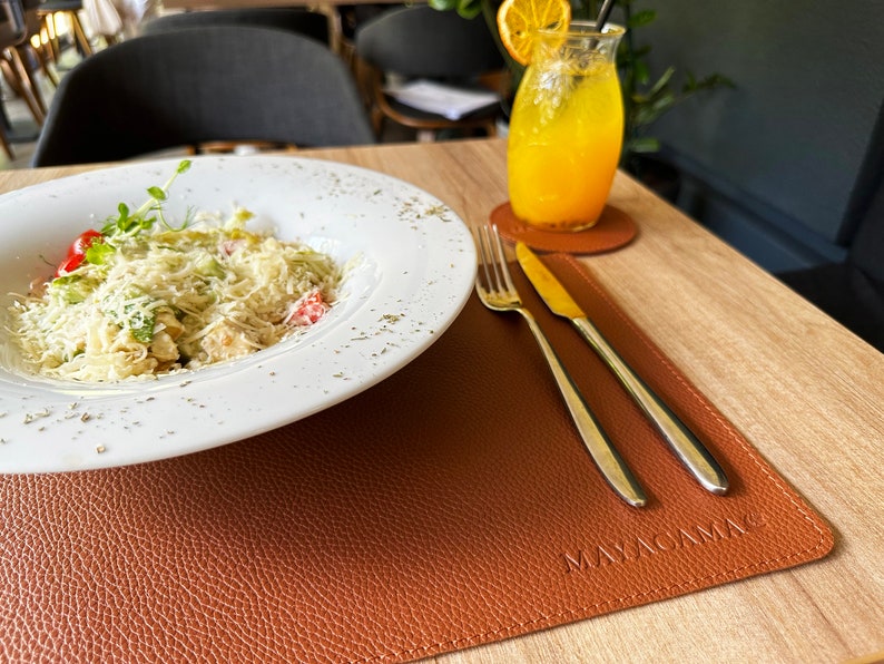 May include: A white plate with a salad topped with grated cheese, tomatoes, and a sprig of greenery. The plate is on a brown leather placemat with the text "MAYACAMA" embossed on it. A fork and knife are on the placemat. A glass of orange juice with a slice of orange is on a coaster to the right of the plate.
