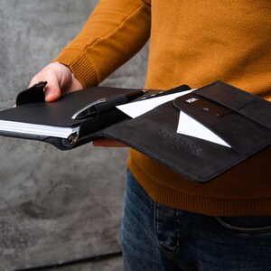 Può includere: Un taccuino in pelle nera con un portapenne e uno slot per carte. Il taccuino è aperto e contiene un foglio di carta bianco.