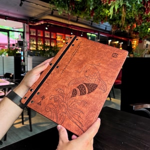 May include: A wooden menu with a koi fish design. The menu is a reddish-brown color and has a black spine with metal rings. The koi fish is etched into the wood, along with Japanese text and wave patterns.