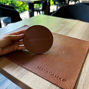 May include: A brown leather coaster with the word "MAYACAMAE" embossed on it. The coaster is on a brown leather placemat with the same word embossed on it.