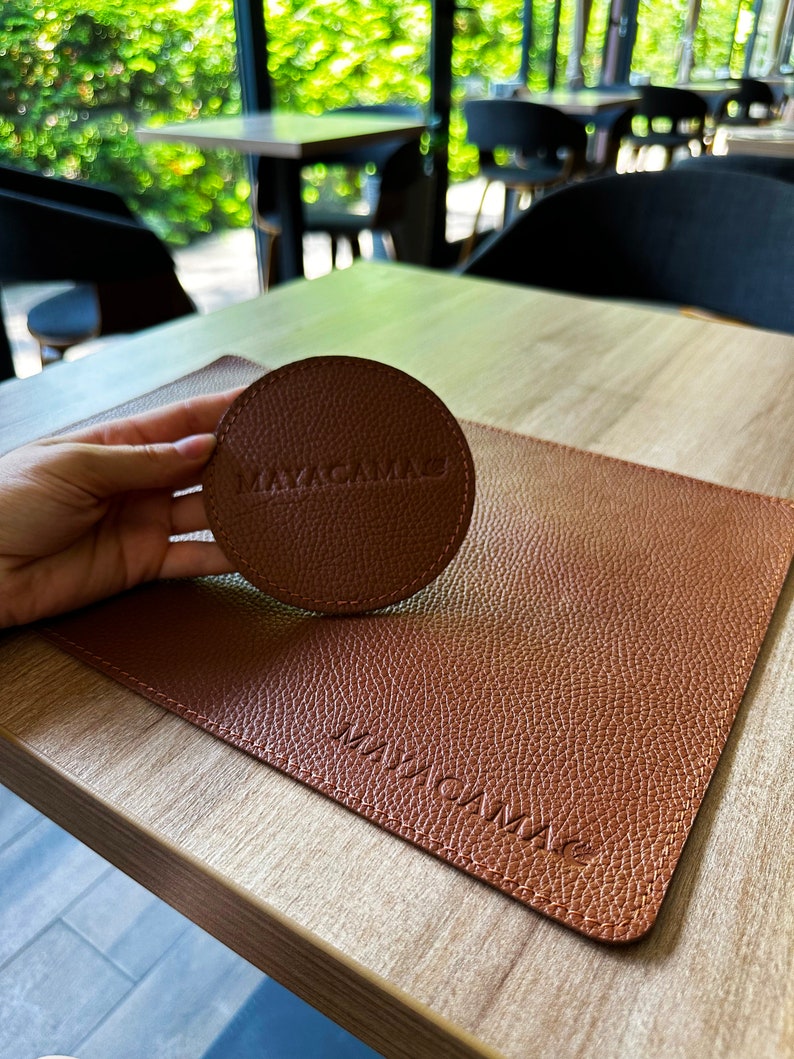 May include: A brown leather coaster with the word "MAYACAMAE" embossed on it. The coaster is on a brown leather placemat with the same word embossed on it.