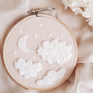 Könnte beinhalten: Ein weißer Stickrahmen mit beigefarbenem Hintergrund, der eine Sichelmond, Sterne und flauschige weiße Wolken zeigt. Der Rahmen ist aus Holz und hat eine natürliche Oberfläche.