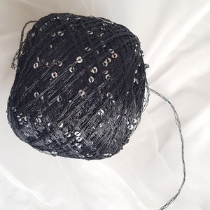 May include: A ball of black yarn with silver sequins woven into the yarn. The yarn is on a white surface and a long strand of the yarn is trailing off to the side.