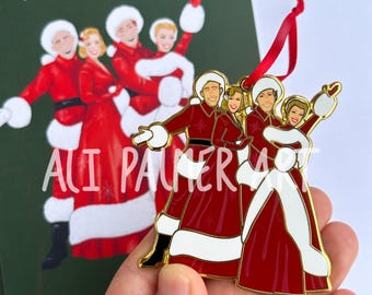 May include: A Christmas-themed ornament featuring a group of people in red and white outfits, with gold trim. The ornament has a red ribbon for hanging. The background shows a matching illustration. The text "ALI PALMER ART" is visible.