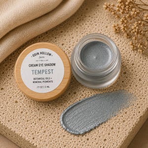 May include: A small glass jar of metallic silver cream eye shadow with a light wood lid. The label reads Odin Hollow Farm, Cream Eye Shadow, Tempest, Botanical Oils + Mineral Pigments, .17 oz | 5 ml. A silver cream swatch is visible nearby.